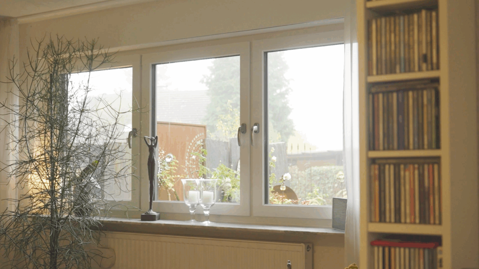 Fenster-2 Gemütliches Wohnzimmer mit großen Fenstern, Zimmerpflanze, CDs im Regal und Kerzenhalter auf der Fensterbank.