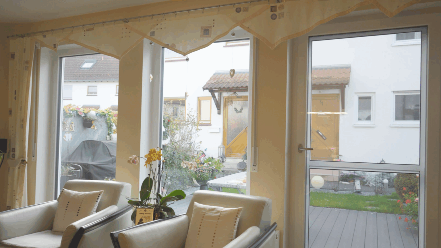 Fenster-21 Gemütliches Wohnzimmer mit großen Fenstern, Blick auf gepflegten Garten mit Pflanzen und überdachter Terrasse.